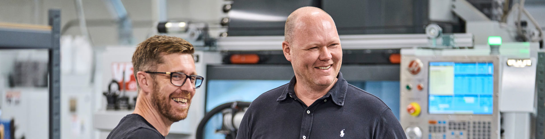 To kolleger fra Induflex står foran en drejebænk og snakker om et plastemne.