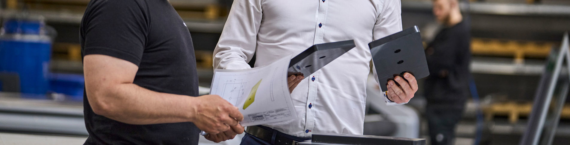 En sælger holder to maskindele i plast op og kigger på en industritekniker, som holder en tegning. 