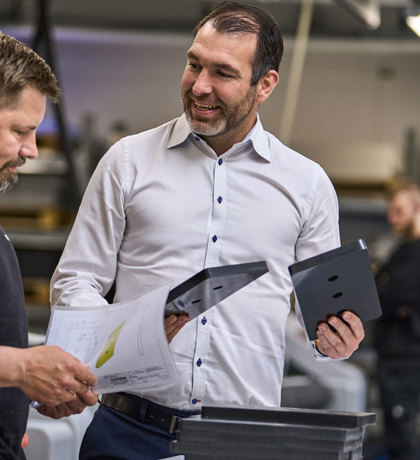 A consultant holds to machine parts in plastics while looking at an industrial technician, which holds a drawing.