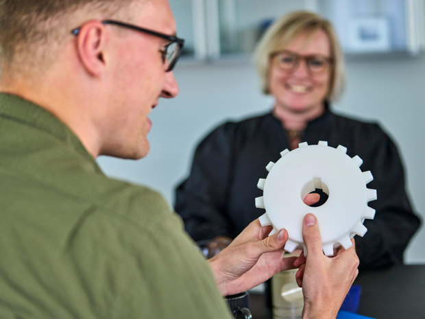 An employee holds a white machine part in his hands during a meeting. 