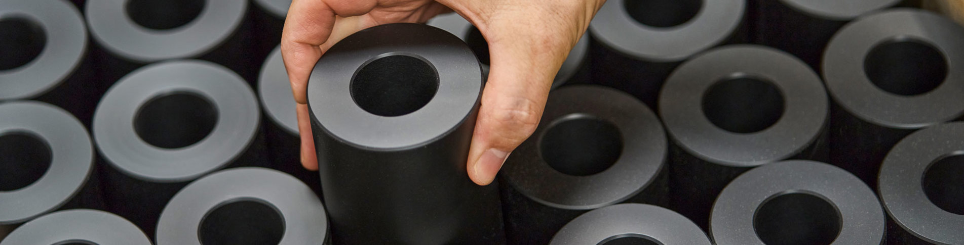 En hånd løfter en drejet maskindel i sort plast fra en palle fyldt med maskindele.