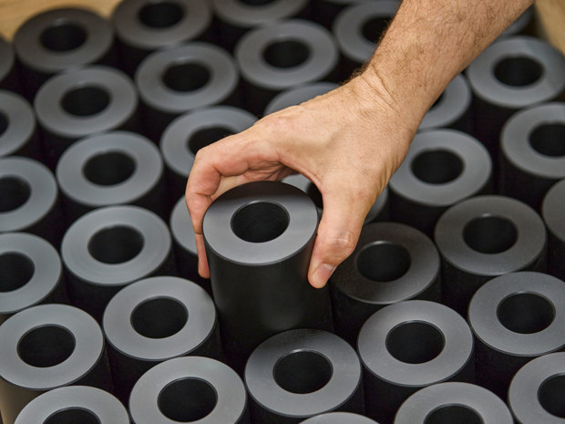 A hand lifts a turned machine part in black plastics from a pallet filled with machine parts.  