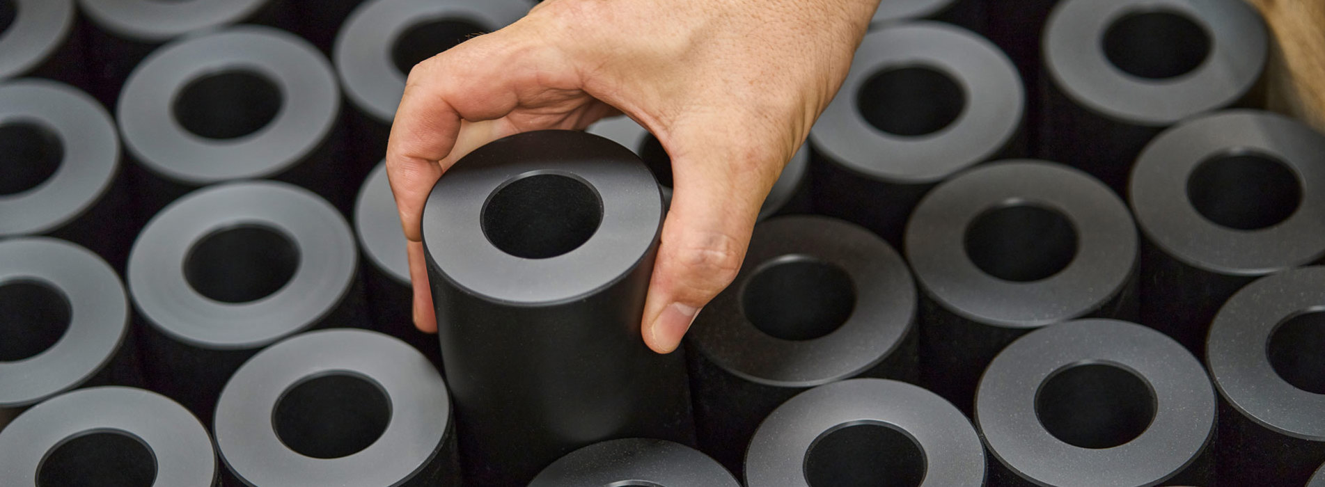 En hånd løfter en drejet maskindel i sort plast fra en palle fyldt med maskindele.