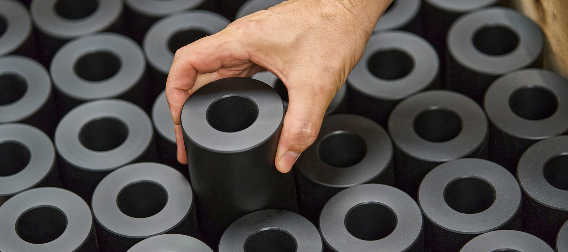 En hånd løfter en drejet maskindel i sort plast fra en palle fyldt med maskindele.